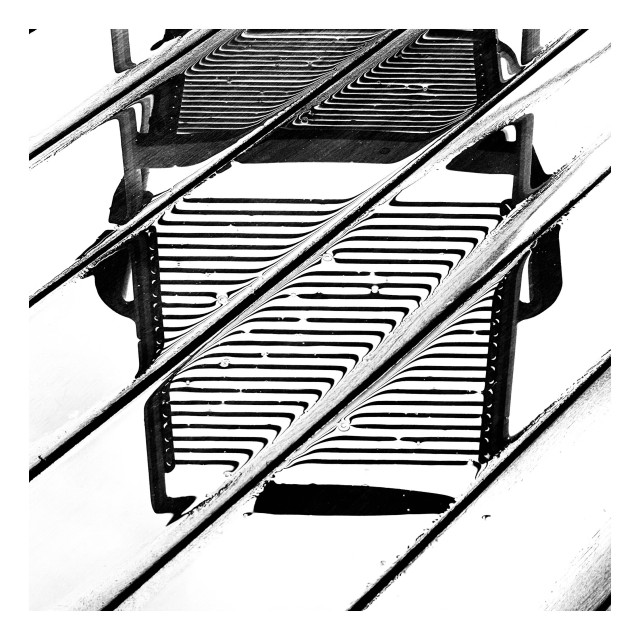High contrast monochrome image of a chair with a corded back and seat reflected in the wet planks of a wood patio. 