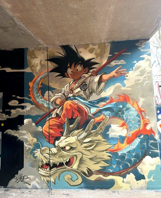 Streetartwall. A cool mural with the manga characters from Dragonball Z was sprayed on a concrete wall under a bridge. It is intended as a tribute to the recently deceased illustrator Akira Toriyama. It depicts an Asian-looking boy (Songoku) with spiky black hair, wearing red trousers and a white kimono shirt and leaping through the clouds with a wooden longstick. Below him flies a fierce-looking blue dragon with red spikes. 
Info: Akira Toriyama (April 5, 1955 - † March 1, 2024) was a Japanese manga artist, character designer and screenwriter. He became known worldwide primarily as the author of the manga series Dragon Ball. Dragon Ball Z is the name of a second television series based directly on the Dragon Ball manga.