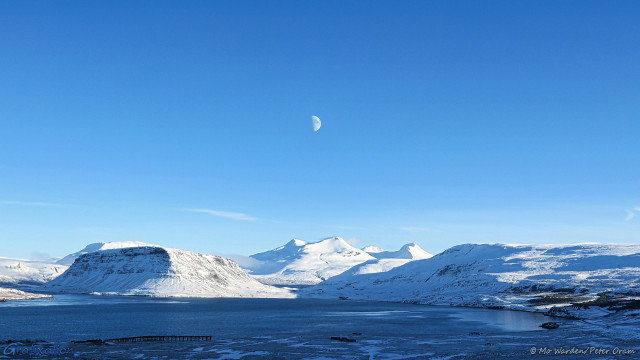 A photo of a fjord beneath a clear blue sky with a few tiny wispy clouds. The water is surrounded by snow- and ice-covered mountains, strongly lit from the right. In the centre of the shot is the moon, almost perfectly half-illuminated, against a cyan background. The mountain directly beneath it has two peaks of nearly equal height, spaced slightly apart and directly below the half-moon, and a ridge line on the right of these peaks which looks like an eye. It's almost as if the mountain is the head of a man lying on his back with his mouth open, attempting to bite the moon.