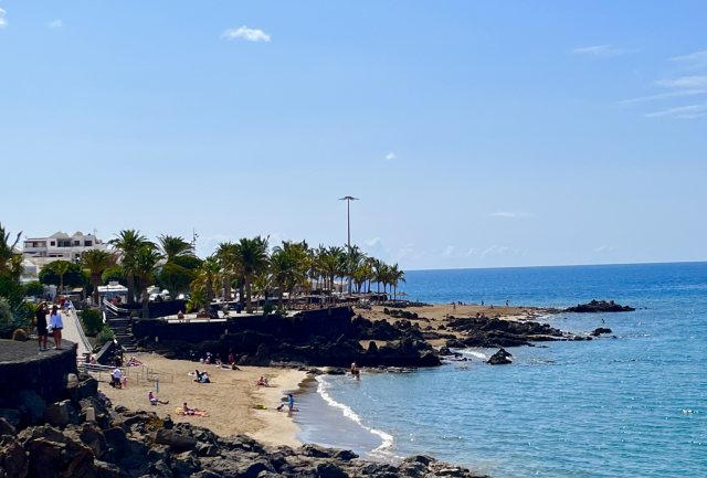 Foto di una parte della costa di Lanzarote, Isole Canarie. Cielo azzurro e terso, mare blu e spiaggia di sabbia fine. Palme a decorare questo spazio di relax. 