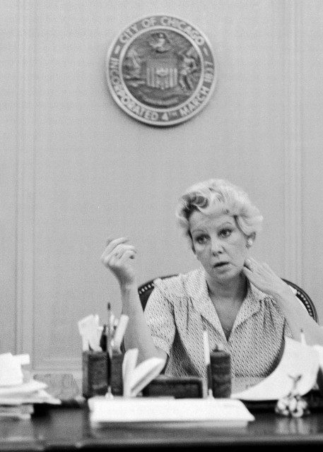 Jane Byrne sat in front of the city of Chicago seal. She is a white woman with blonde hair.