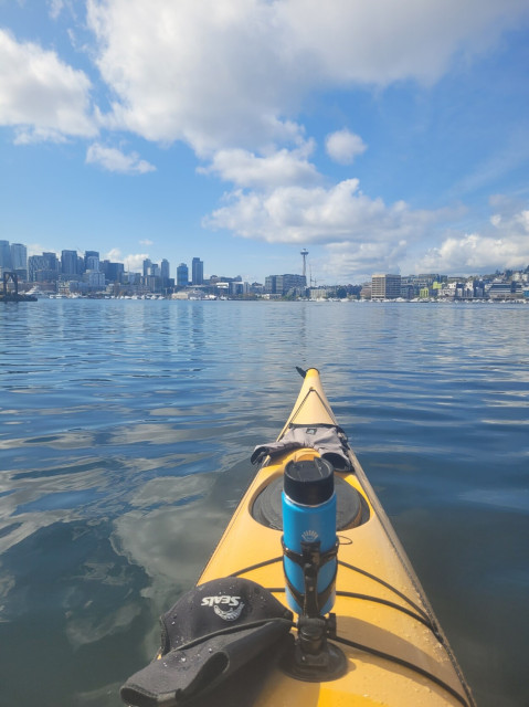 Front of yellow kayak in a calm lake with downtown Seattle skyline in background. Sunny skies, fluffy white clouds.