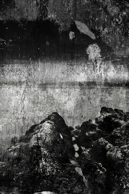 Black and white photo of craggy, textured rocks, with the sea beyond. With a textured overlay of slate and rolled ink.