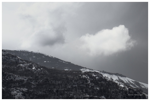Low hanging white cloud in front of a background of gray rain clouds. There are diagonal, snow covered forest hills. The composition vaguely resembles the yin yang symbol.