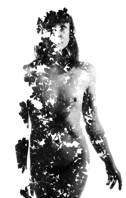 A naked woman is standing in front of the camera. Her silhouette is filled with holes from the double exposed leaves through the trees. Black and white. 