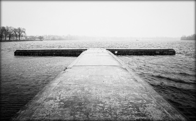 Looking down a cement t-shaped wharf on a small lake. Choppy water and a pure white sky are seen, the result of a late winter storm.