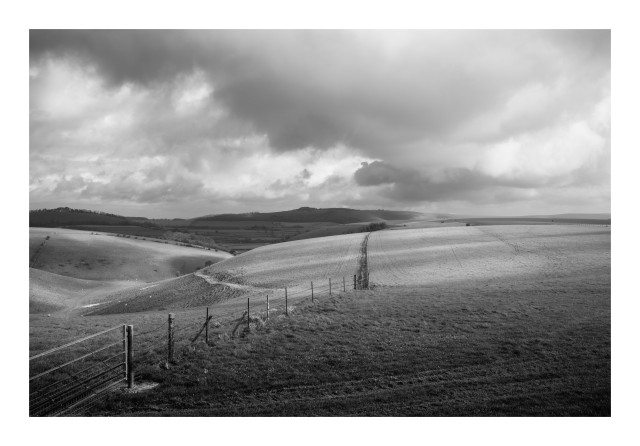 A black and white image of a countryside scene. There are rolling fields and hills, with large voluminous clouds, and a fence taking you into the image.