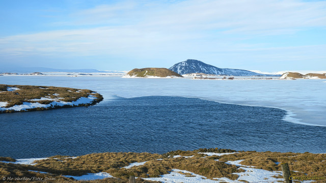 A photo of a landscape with snow and ice. The foreground is brown vegetation with patches of snow, and the horizon line is what appears to be islands with a mountain beyond. The sky is clear cyan but with a fair amount of wispy cloud. The centre of the shot is filled by a lake, reflecting the blue of the sky where it's liquid, and with a coating of ice further from the shoreline. The low sunlight is from the left.