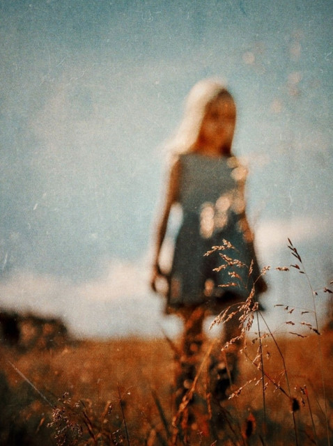 Photography. A color photo of a little girl in a summer meadow. The picture is deliberately blurred. You can't make out the blonde girl running across the wide orange meadow in her blue dress. You can make out the blue sky, the wide landscape and a few dark trees at the edge. But the focus is on a few blades of bright grass in the foreground. A warm and calm photo that conveys a feeling of past summers and beautiful memories.