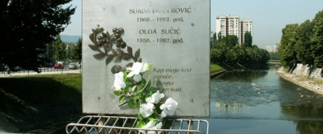 A photo of the memorial sign on the bridge. Someone has placed white flowers in the basket designed for that. Photo by jaimesilva on flickr.