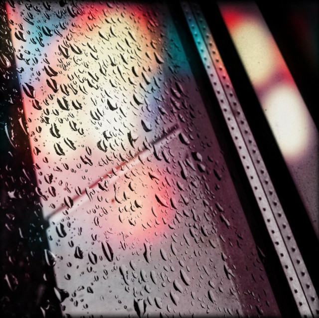 A rain dropped covered window is shown with different colours being formed from nearby lights.