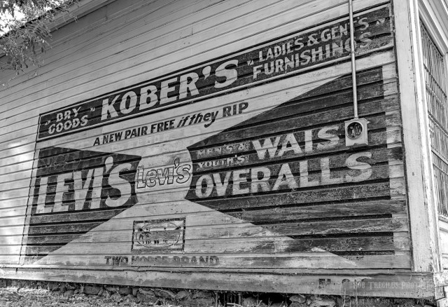 Photo in black and white of a painted sign on the side of an old wood plank building that reads: "Dry Goods. Kober's. Ladies & Gent's Furnishings. A new Pair if they RIP. Levi's. Levi's. Men & Youth's Waist Overalls. Two Horse Brand."