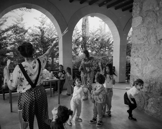 Black and white image: Under a stone colonnade in a garden, two clown-costumed adults entertain a group of young children by blowing and releasing soap bubbles. The kids’ faces reflect their delight.