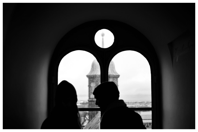 Schwarz-Weiß-Foto. Blick durch ein Fenster mit zwei Rundbögen und einem kreisrunden Fenster. Dahinter eine Kirche und Häuser von oben, vor dem Fenster zwei Menschen als Silhouette. 