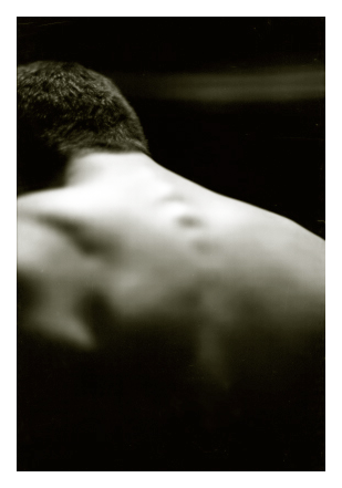 lit by by bare bulb. close up. a white guy in his 20s with short, dark hair leans forward and left. muscles in his back and his spine are defined. the background is black with a faint horizontal brush of gray.
