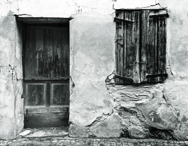 Black and white photo of an abandoned house in Serralunga, Piedmont. You can see a cracked wall, an old locked door and to the right of it a barred window whose shutters are just as weathered as the front door. Nobody lives here anymore.