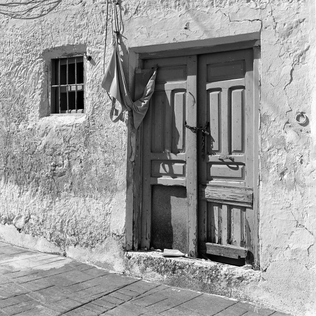 A black-and-white photo of an old wooden front door. The door is secured with a chain and padlock. Some timber door panels are missing.