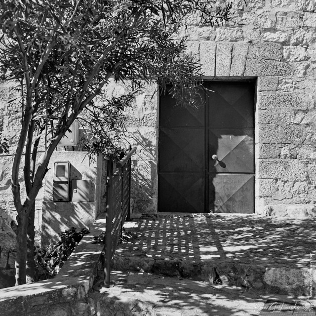 A black-and-white photo of a metal door round the side of the church. The church wall is stone and there is a young tree growing on the LHS.