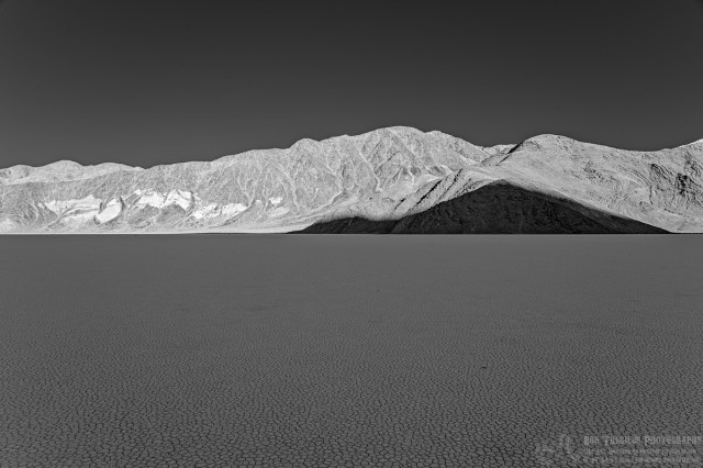 Black and white landscape photo of a dry lake bed. The foreground dry playa is checked with polygonal mud cracks. The background are bare rugged mountains. The sky is nearly black. There is a triangular shape shadow on the mountain range cast by another mountain behind the photographer in this late afternoon photo.