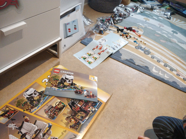 The box for a Lego set is in the foreground, with images of the various builds that can be made. one of the instructions are on top of the box, showing a Skull Island build. 

In the back is an open instruction book with the different bags of Lego pieces laid out in order. One bag has been opened with it's contents laid out next to the book. 

A child's knee and foot been be seen on the right side of the photo.