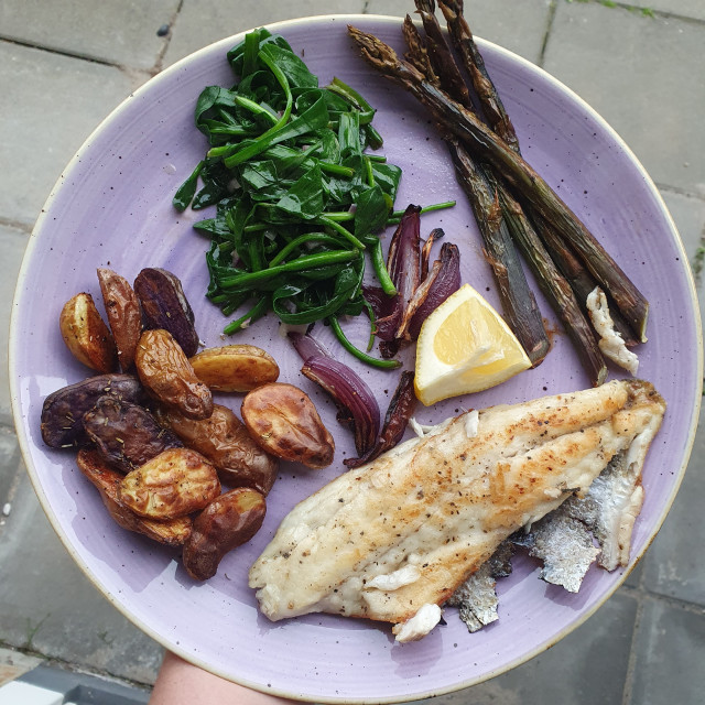 A purple plate with a pan fried fish filet surrounded by sides: toasted purple asparagus, purple (&white) potatoes and red onions and sea lavender (a kind of small green leaves, cooked). 