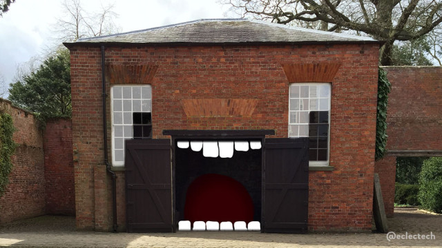 Photo of a large red brick outbuilding, somewhere in Yorkshire. It has large double garage doors, wide open, exposing a dark interior. Up and either side of the doorway are tall sashed windows, with bricks above them arranged vertically. It has a shallow peaked roof and you can see large trees behind. I have tweaked the picture, so the eye-like nature of the windows is emphasised by darkening the bottom right into a rectangular pupil, and making the rest lighter. I have added the hint of mouth interior inside the building, and teeth along the top and bottom. It looks quite excited about something.