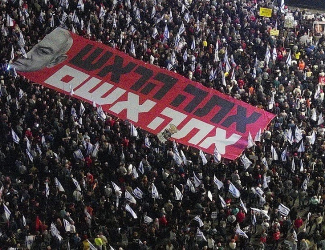 A large crowd with a banner featuring the  face of Netanyahu and text saying: you’re the head (prime minister), it’s your fault 