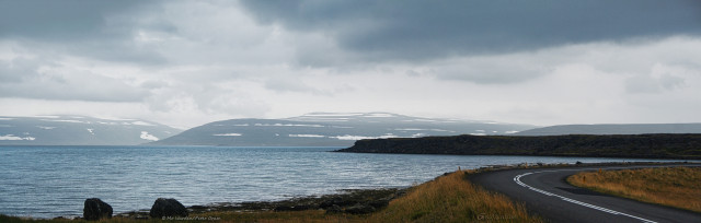 A panoramic photo of sea and mountains. On the right is a tarmac road, surrounded by brown grassland, leading down to a gravelly beach. In the centre and left is a rippled fjord. In the centre is another promontory and, on the horizon, a line of misty mountains with lines of snow along their flanks. A deep cleft is visible in the mountains, and above the whole scene is a cloudy sky with a heavy grey bank of raincloud overhead.