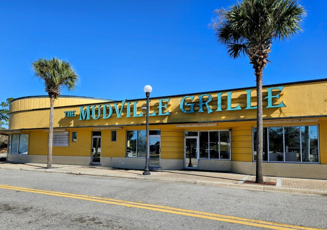 Popular sports bar and restaurant in a long old fashioned looking building painted bright yellow with large teal colored signage spelling out Muddville Grille. With palm trees framing the building all beneath a clear blue sky.