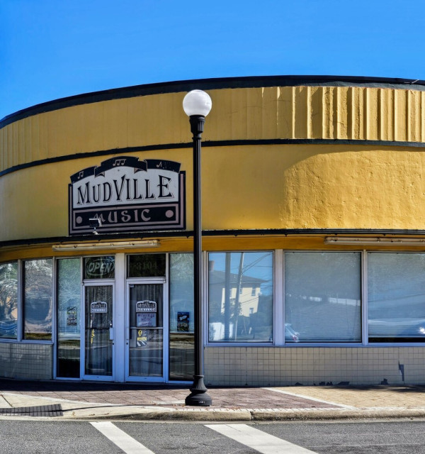 The rounded corner of an old building painted bright yellow with signage in black for the "Muddville Music." Inside is a popular music room with live big band music.