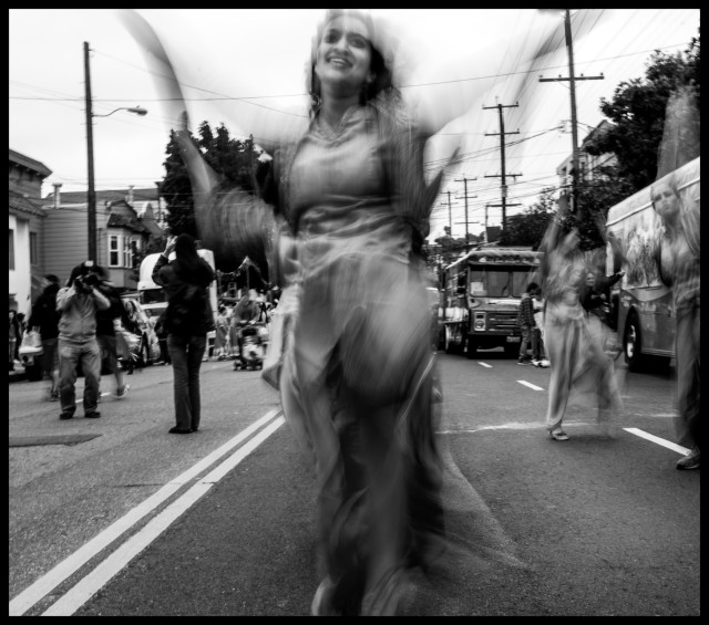 A monochrome photo of a woman celebrating Carnaval, dancing in the street. Taken with a 1/6th second exposure, she is a blur of motion... yet well enough defined that you can see her arms have just been raised up like wings, about to take flight. Her face is caught in glorious rapture. 
