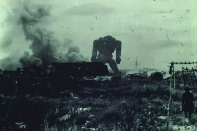 A human-shaped mech looms over a burning farm building