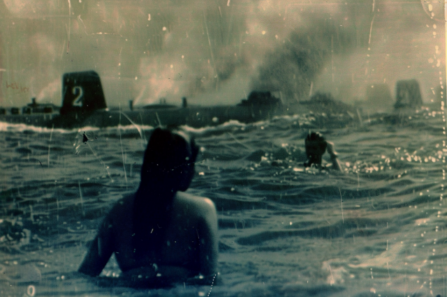 Swimmers gaze in shock at the submarines which have surfaced just offshore. Clouds of smoke billow in the distance