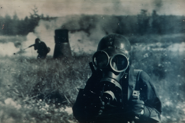 A soldier in chemical warfare suit and mask faces the camera. Behind another soldier aims their rifle at something unseen in long grass