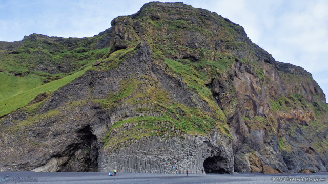 A photo of a mountain face, with grey rock and green grass slopes. At the foot of the mountain is a grey, powdery beach, this is actually the sand which is black when wet. The middle of the rock face is comprised of vertical columns of basalt resembling an elephant's trunk and, on either side, there are two caves looking like the eye sockets on an elephant's head. There are a few people at the base of the cliff, taking photos.