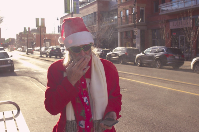 A man dressed as Santa Claus holds his phone while smoking marijuana. He is wearing sunglasses. Standing on the sidewalk near a bus stop. Sunny winter day, no snow tho.
