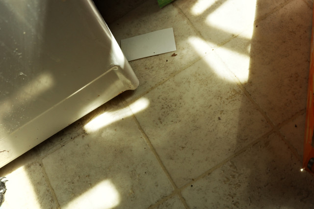 gr iiix view of shadows on the tile floor + the corner of a washer + a white card sticking out from underneath it + the very edge of an orange box + the light from the window making shiny patches