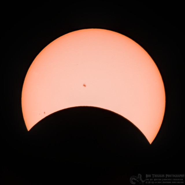 A color photo of the sun partial obscured by the moon. It looks like someone took a bite out of the sun and a third of it is missing. There's two prominent sunspots: one at center and the other on the lower left of the sun's disk.
