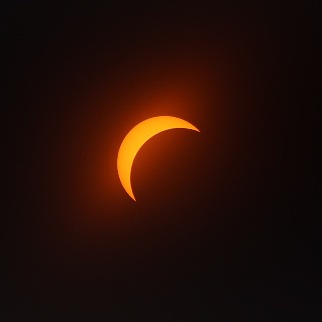 Photograph of the orange, crescent sun through a filter as the moon passes in front of it, covering about 70% of it from the lower right.