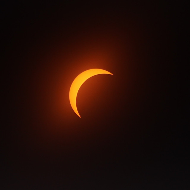 Photograph of the orange, crescent sun through a filter as the moon passes in front of it, covering about 80% of it from the lower right.