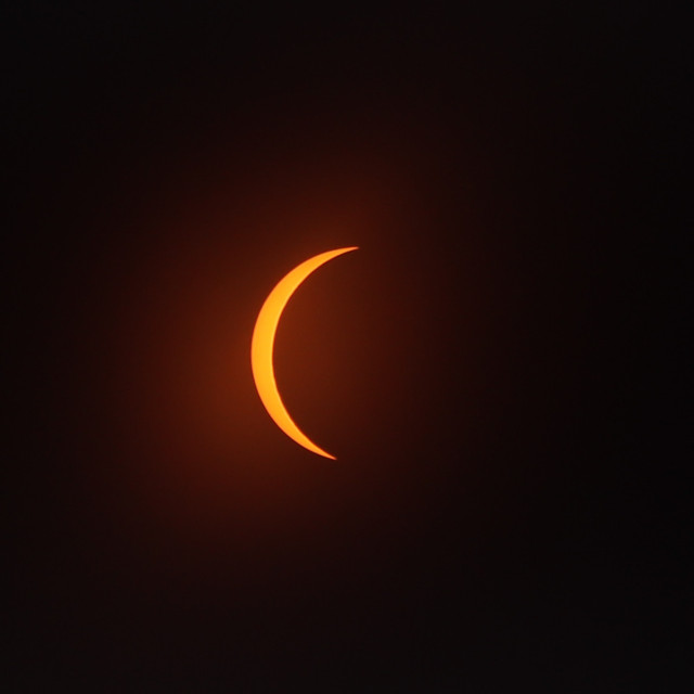 Photograph of the orange, crescent sun through a filter as the moon passes in front of it, covering about 90% of it but leaving the left side of the sun exposed.