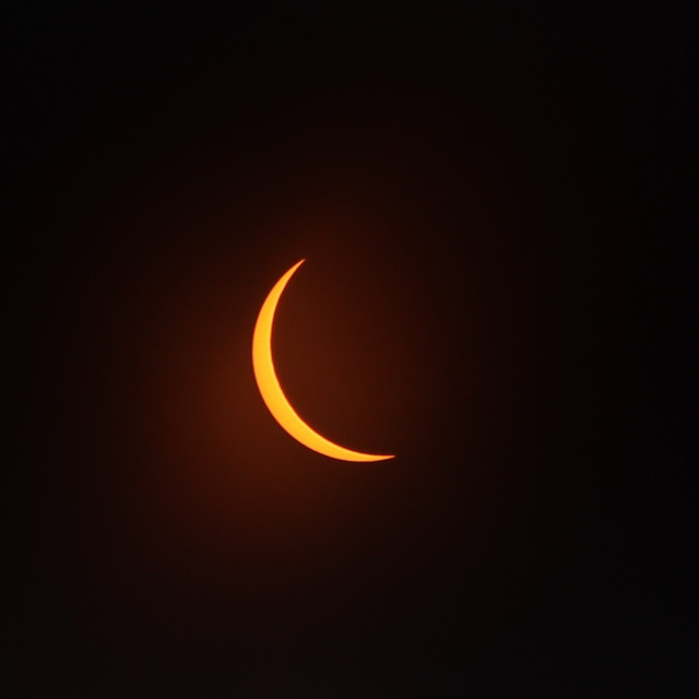 Photograph of the orange, crescent sun through a filter as the moon passes in front of it, covering about 90% of it with the left side of the sun exposed and the top getting covered as the bottom edge emerges.