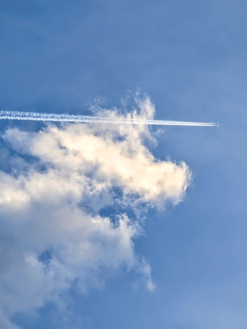 A photo of the blue sky with white clouds and a jet on the right side leaving a streak across the sky.