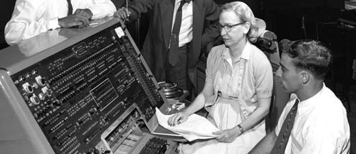 Grace Hopper at the controls of a computer, surrounded by white men in ties. She is a white woman with light hair.
