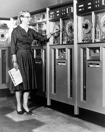 Grace Hopper with a bank of mainframes and the COBOL manual. She is a white woman with light hair.
