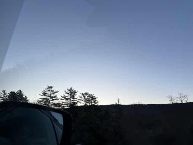 A view out the car window. You can see the silhouette of a mountain with the light colors of dawn behind it. A few nearby trees poke above the Ridgeline of the mountains. Mirror to the car. You can see a pond with morning sunlight, reflecting off of it. There is a guard rail with heavy motion, blur, and the car mirror.