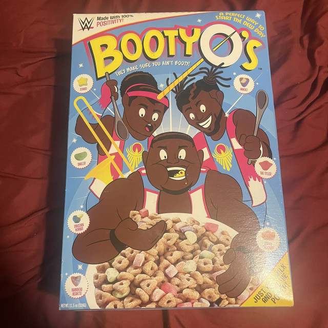 A  playful and colorful cereal box for a product called "Booty O's," with a tagline that reads "They make sure you ain't booty." The box art is lively and cartoonish, featuring three animated characters who are joyfully presenting the cereal. The characters appear to be wrestlers, and their attire includes wrestling boots and vibrant, superhero-like costumes. They are surrounded by various symbols like stars, rainbows, and unicorns, which add to the whimsical and energetic feel of the design.

The cereal itself consists of oat pieces shaped like stars, unicorns, and rainbows, suggesting a fun and magical eating experience. The box includes a note that it's "A perfect way to start the New Day," possibly the New Day group in the WWE entertainment  universe. The overall design conveys positivity and excitement, and is likely to attract those who enjoy playful branding and are fans of vibrant, character-themed products.