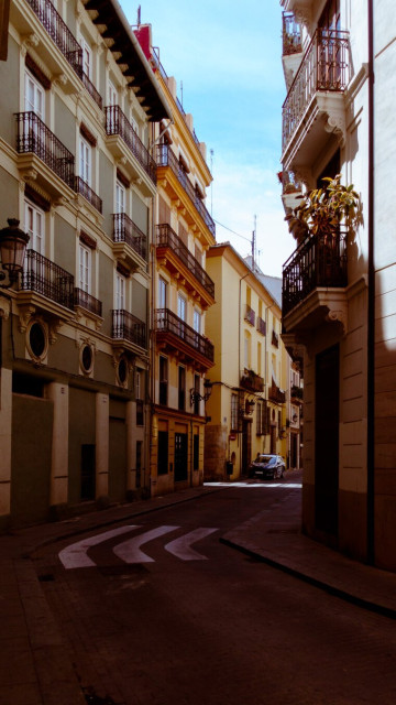 Shot of a street in the historic city center of Valencia. The street has a slight curve to the right and a bit further down there is a police car standing. The street is mostly in the shadow with the exception of a light ray right in front of the police car. The colours are warm.