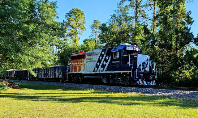 Led by a red-white-and-blue painted engine, a train passes by a grassy field with a tall treeline on the opposite side, behind the train. The engine has markings from Florida East Coast Railway and parent company, GrupoMéxico, and a yellow ribbon below the words: Honoring our Veterans.  The engine pulls several rust-brown colored open-top train cars with colorful graffiti on their sides, filled with a light colored sand or gravel.