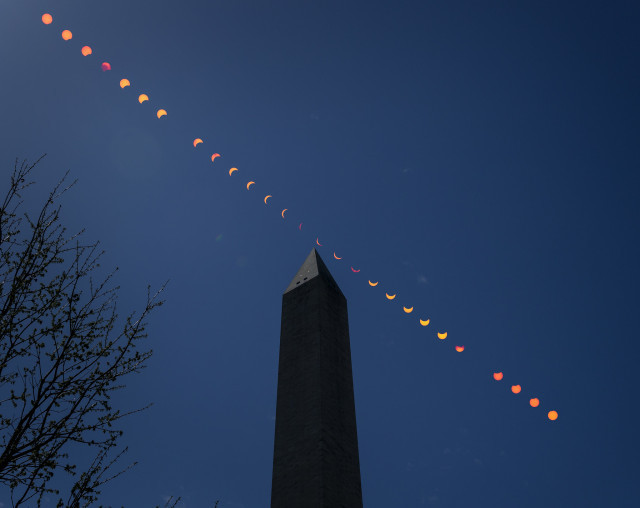 This composite image shows the progression of a partial solar eclipse over the Washington Monument in Washington. The orange Sun and lunar shadow track from the upper left to the lower right of the frame, across a dark blue sky over the pointed tip of the silhouetted obelisk. The bare branches of a tree reach into the lower left side of the frame. Credit: NASA/Bill Ingalls⁣